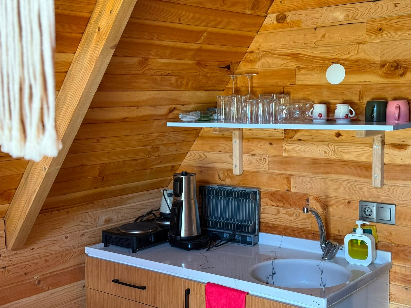 Kitchenette with a kettle, hob, sink and a shelf of glassware against the pine wall.